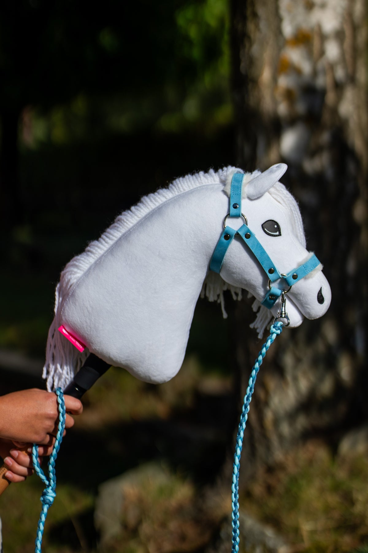 Detailfoto: de kop van Hobby Horse Lady met zwart Halster van Hobby Horse LarDen wordt omhooggehouden in gevlekte schaduw – wit pluche, donker oog, witte manen van garen – hier getoond met het Pastell Blue-bonthalster van kunstleer en het Double Blue-gebreide halstertouw