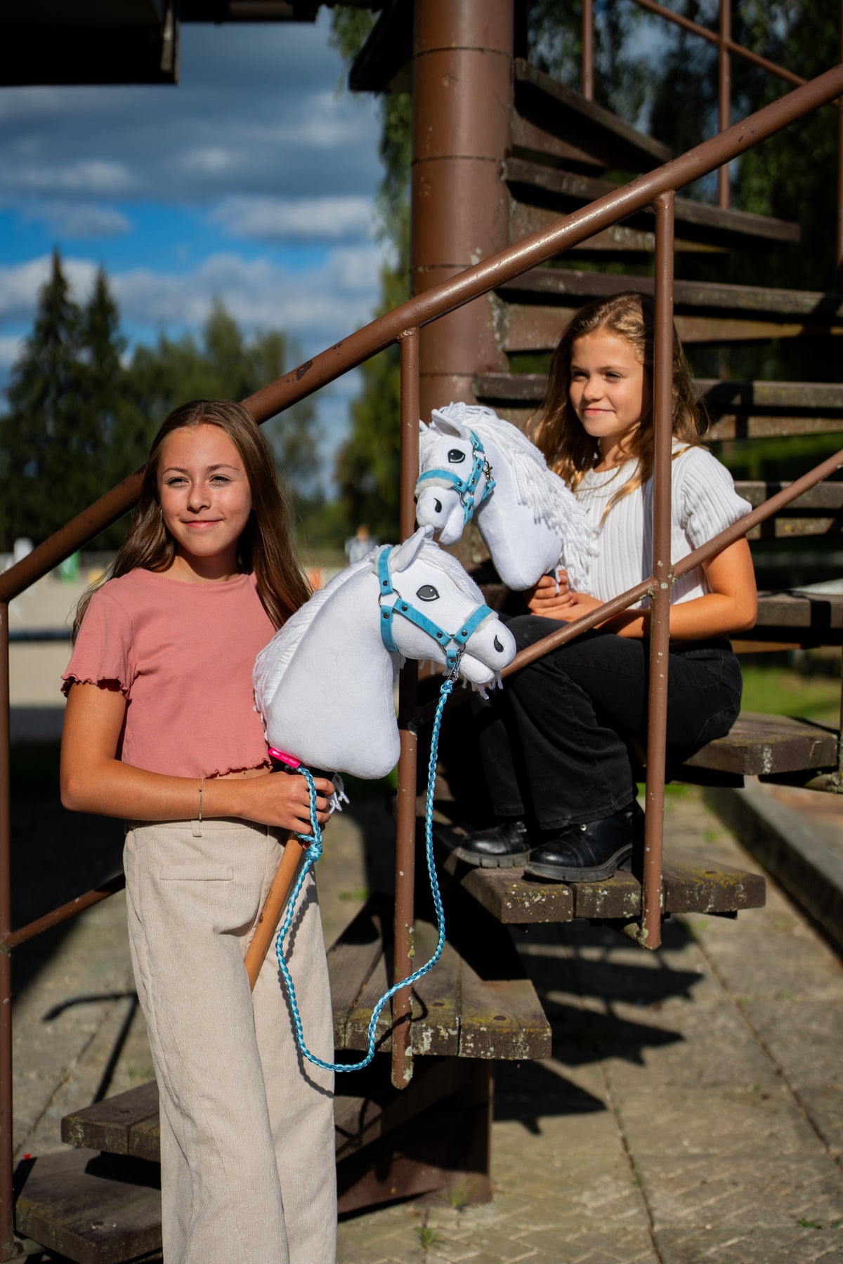 Lifestylefoto: twee meisjes met witte Hobby Horses van Hobby Horse LarDen op een roestige metalen trap, met Hobby Horse Lady met zwart Halster vooraan – witte pluchen kop, witte manen van garen – hier getoond met het Pastell Blue-bonthalster van kunstleer en het Double Blue-gebreide halstertouw