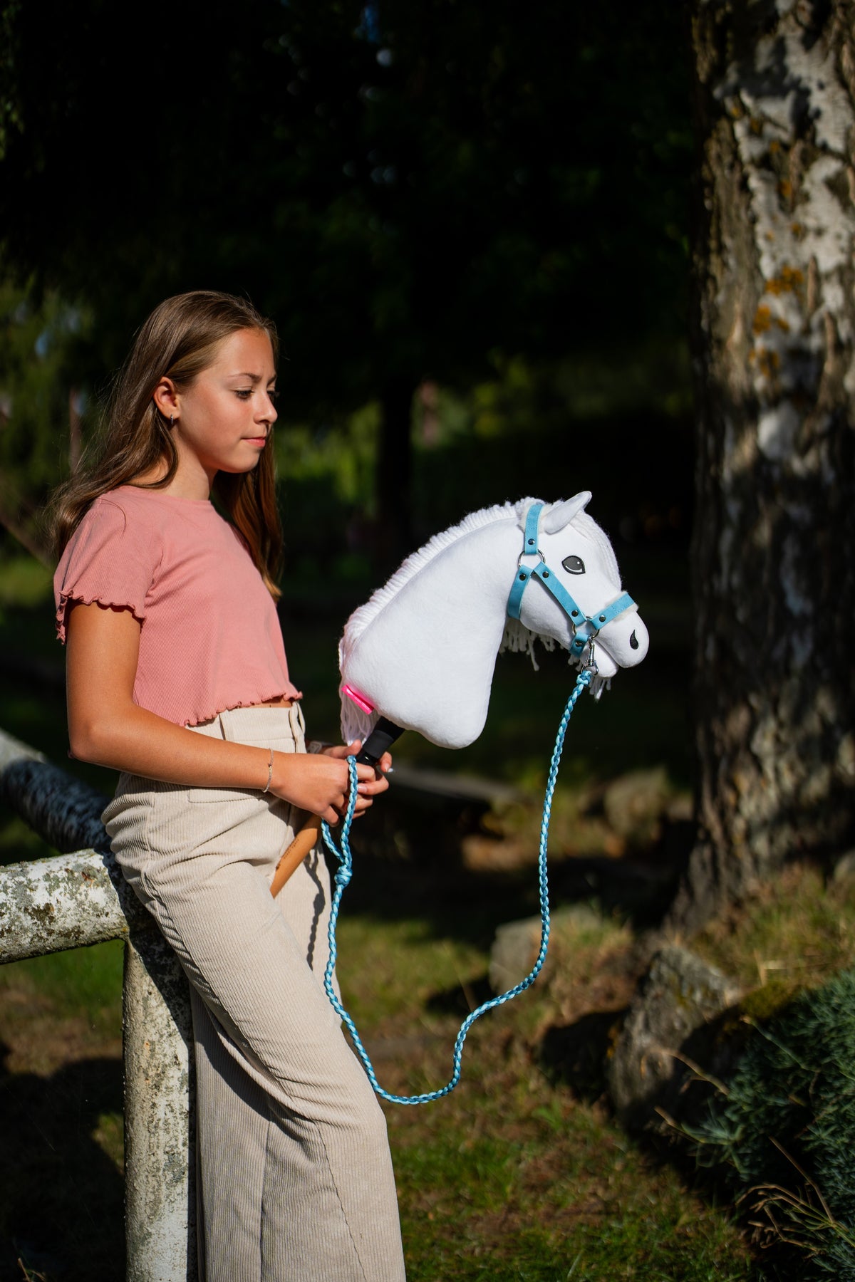 Lifestylefoto: een meisje in een roze top staat bij een bemoste boom met Hobby Horse Lady met zwart Halster van Hobby Horse LarDen, met het halstertouw in een lus hangend – witte pluchen kop, witte manen van garen – hier getoond met het Pastell Blue-bonthalster van kunstleer en het Double Blue-gebreide halstertouw