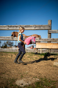Een meisje in rijkleding loopt over een zanderige paddock met de Hobby Horse LarDen Pinkie met glitterhalster van Hobby Horse LarDen - roze pluche hoofd met roze-witte wollen manen - hier getoond met de Halster turquoise