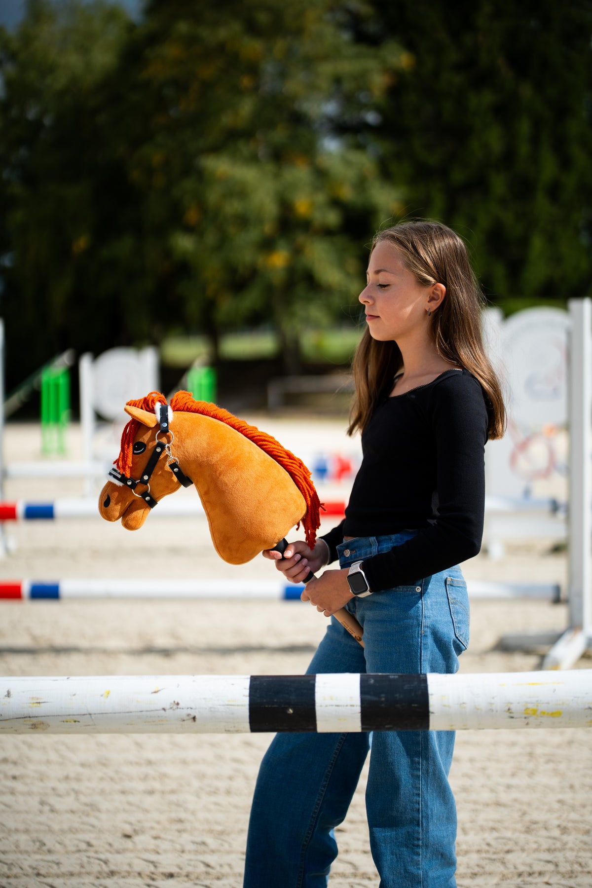 Lifestylefoto: een meisje in een zwarte top staat achter een springbalk op een zandpiste en houdt de Hobby Horse Little Angel met zwart Halster van Hobby Horse LarDen vast - voskleurige pluchen kop, felrode garenmane - hier getoond met het Espresso bonthalster van kunstleer