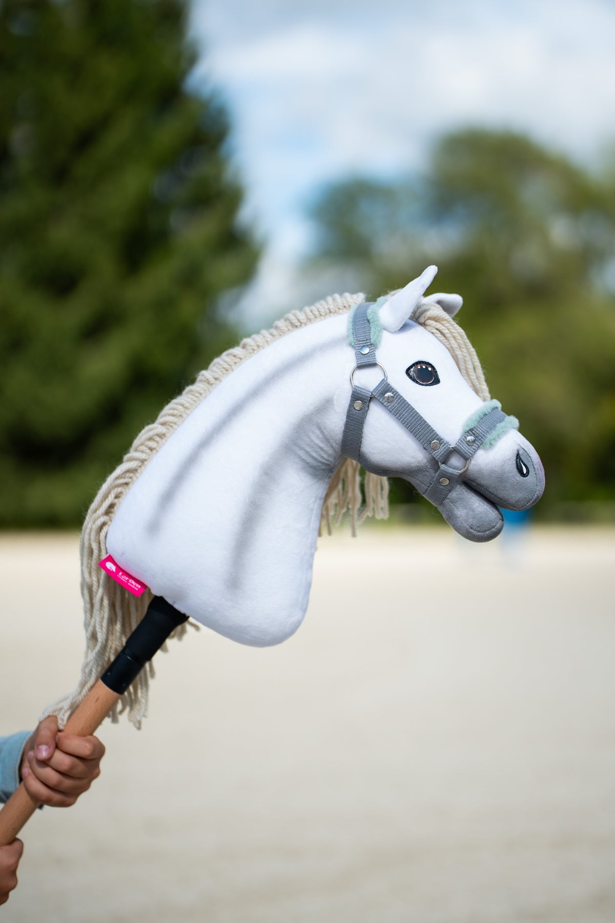 Detailfoto: de kop van Hobby Horse Little Lady met zwart Halster van Hobby Horse LarDen wordt omhooggehouden op een zandrijbaan, met coniferen erachter – wit pluche, crèmekleurige manen van garen en voorpluk – hier getoond met het grijze halster met mintgroen bont