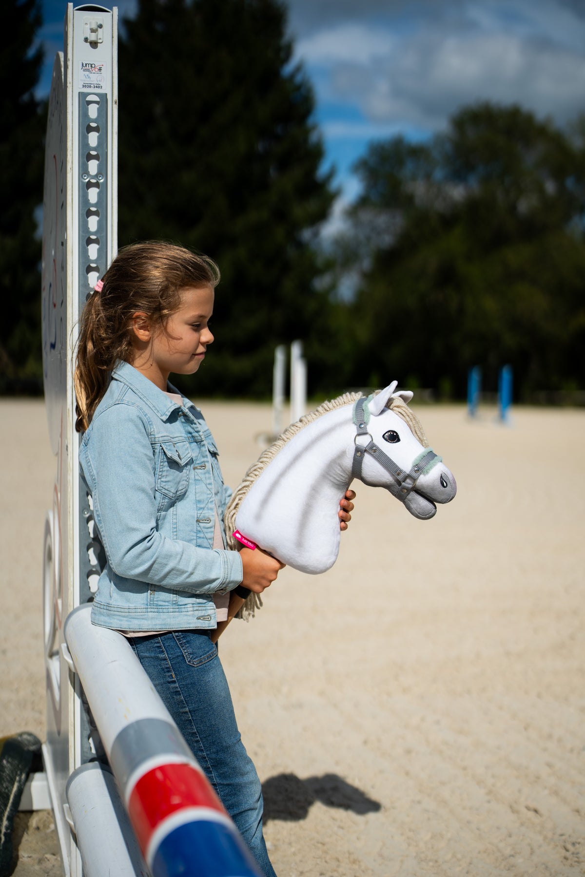 Lifestylefoto: een meisje in een spijkerjasje staat bij een hindernisstaander op een zandpiste en houdt de Hobby Horse Little Lady met zwart Halster van Hobby Horse LarDen vast - witte pluchen kop, crèmekleurige garenmane - hier getoond met het grijze Halster met mintkleurig bont