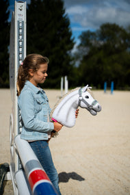 Lifestylefoto: een meisje in een spijkerjasje staat bij een hindernisstaander op een zandpiste en houdt de Hobby Horse Little Lady met zwart Halster van Hobby Horse LarDen vast - witte pluchen kop, crèmekleurige garenmane - hier getoond met het grijze Halster met mintkleurig bont