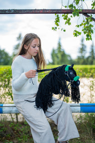 Lifestylefoto: een meisje in een lichte trui zit op een paddockhek en verzamelt de teugels van Hobby Horse Maximus met zwart Halster van Hobby Horse LarDen – zwarte pluchen kop, lange zwarte manen van garen – hier getoond met het zwarte kunstleren halster met groen bont en zwarte kunstleren teugels
