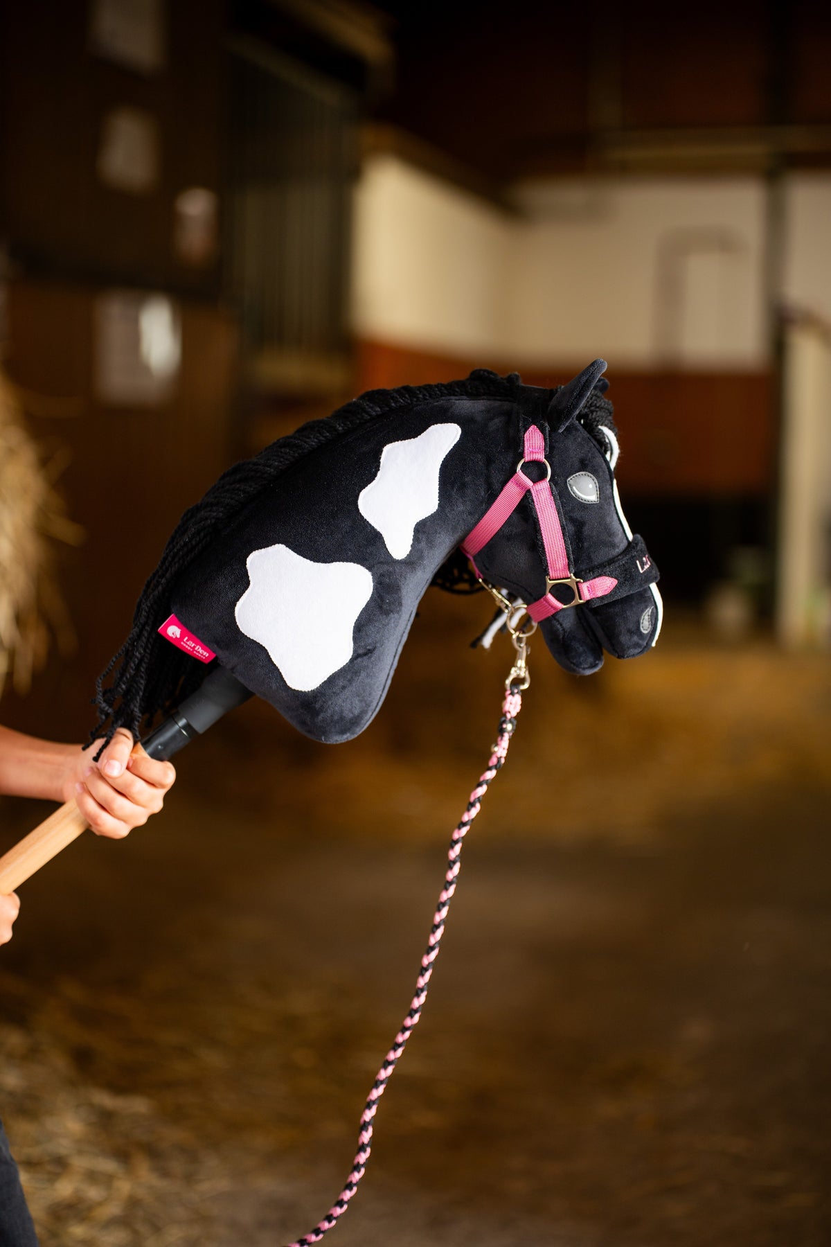 Het hoofd en de hals van het Hobby Horse Merlin met Black Halter van Hobby Horse LarDen worden in een stal omhooggehouden, met zwart pluche en witte vlekken en zwarte manen van garen, hier getoond met de LarDen Exclusive Pink-set en het gevlochten roze halstertouw