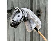 A close-up of the Hobby Horse LarDen "Hobby Horse Naivy with Black Halter," a gray Appaloosa stallion hobby horse featuring a white yarn mane and black spots, posed before a weathered wooden door.