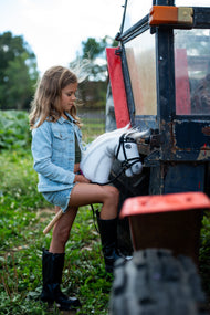 Lifestylefoto: een meisje in een spijkerjas zet haar laars op de trede van een rode en zwarte tractor, met Hobby Horse Nexus met zwart Halster van Hobby Horse LarDen – witte pluchen kop, witte manen van garen – hier getoond met het complete De Luxe-hoofdstel met twee frontriemen