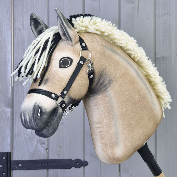 A close-up of Hobby Horse LarDen's beige Hobby Horse Ozzy with a black and white yarn mane, black halter, and button eyes, mounted on a stick against a gray wooden background.