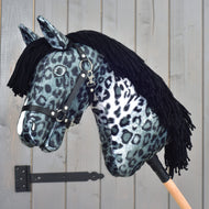 Hobby Horse Picasso met zwart halster van Hobby Horse LarDen, gestyled als een opvallende hengst met zwart-witte stof in luipaardpatroon dat doet denken aan de gevlekte vacht van een Appaloosa, met zwarte manen van garen en een zwart halster, gemonteerd op een houten stok voor een grijze houten deur.