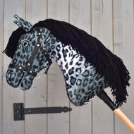 Hobby Horse Picasso met zwart halster van Hobby Horse LarDen, met een pluche hoofd met een grijs-zwarte gevlekte vacht geïnspireerd op een Appaloosa, een zwart halster en zwarte manen van garen, gemonteerd op een houten stok en getoond tegen een grijze houten wand.