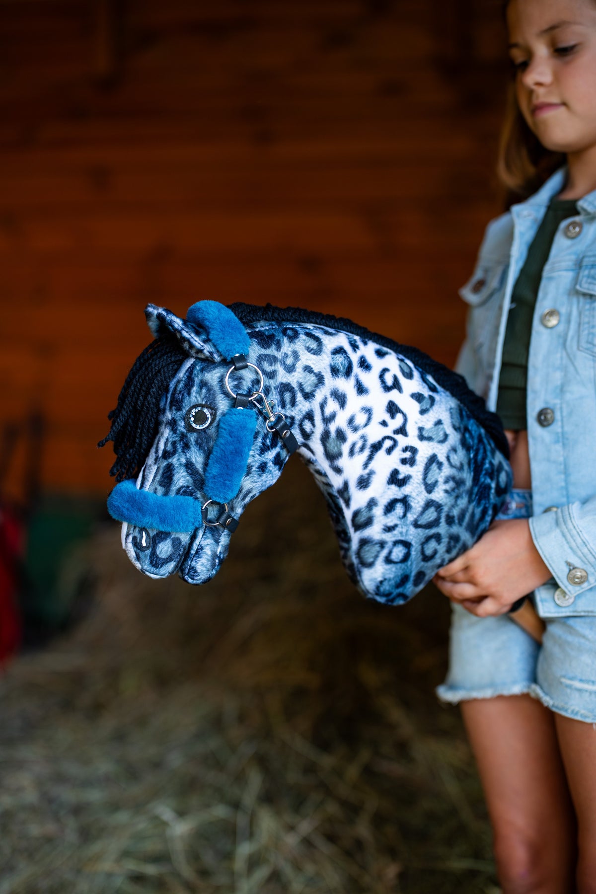 Lifestylefoto: een meisje in een spijkerjas houdt Hobby Horse Picasso met zwart Halster van Hobby Horse LarDen tegen een rode houten schuurwand – blauwe en witte pluchen kop met luipaardprint, zwarte manen van garen – hier getoond met het met bont gevoerde Teddy Petrol-halster