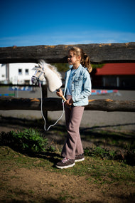 Een meisje in een licht spijkerjasje en een mauve broek staat bij een houten weidehek met de Hobby Horse Pixie met zwarte halster van Hobby Horse LarDen - beige pluche hoofd met witte wollen manen - hier getoond met de Halster grijs met roze bont en het Halstertouw streep Tizian