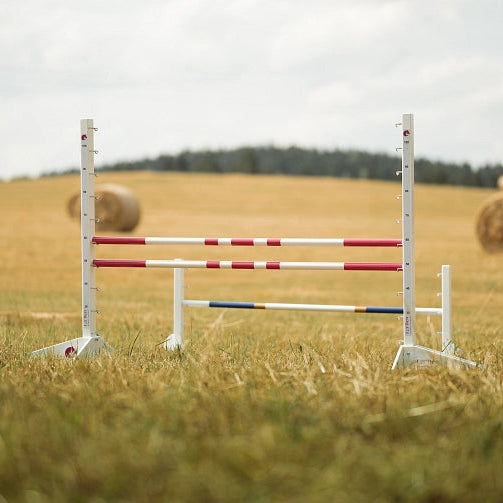 White and red-striped horse jumping poles set up on a grassy field, perfect for Hobby Horsing, with hay bales and trees visible in the background under a cloudy sky.