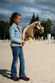 Lifestylefoto: een meisje in een spijkerjas staat op een zandrijbaan met Hobby Horse Smart met zwart Halster van Hobby Horse LarDen, met hindernispalen erachter – palomino pluchen kop, lange crèmekleurige manen van garen – hier getoond met het zwarte kunstleren halster met volledig bont