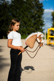 Lifestylefoto: een meisje in een witte top loopt met Hobby Horse Star met zwart Halster van Hobby Horse LarDen over een zandrijbaan, met een gele hindernisvleugel erachter – palomino pluchen kop, golvende witte manen van garen – hier getoond met het bruine halster met volledig bont en de Vivaldi-teugels