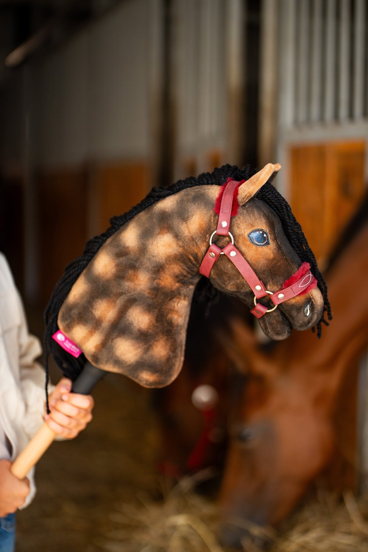 Detailfoto: de kop van Hobby Horse Stormy met zwart Halster van Hobby Horse LarDen wordt omhooggehouden in een stal, met een echt bruin paard wazig erachter – bruin gevlekt pluche, blauw oog, zwarte manen van garen – hier getoond met het Burgundy-bonthalster van kunstleer