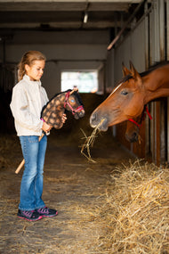Lifestylefoto: een meisje in een crèmekleurige jas houdt Hobby Horse Stormy met zwart Halster van Hobby Horse LarDen bij een echt bruin paard dat hooi eet in een stal – bruin gevlekte pluchen kop, zwarte manen van garen – hier getoond met het Burgundy-bonthalster van kunstleer