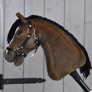 Hobby Horse Sundance met zwart halster van Hobby Horse LarDen met een pluchen ontwerp, zwarte manen van garen, hoofdstel en teugels, dat de levendige uitstraling van een Engelse volbloedhengst vastlegt, gemonteerd op een stok en getoond tegen een lichte houten plankwand.