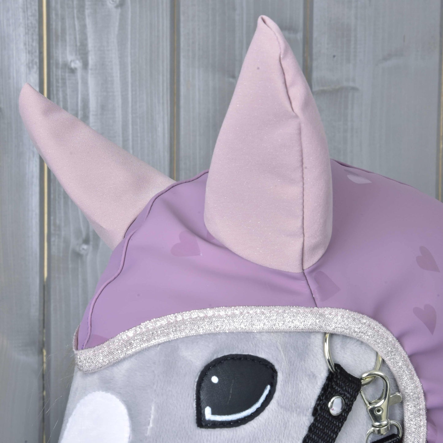 Close-up van de pluchen paardenkop Hobby Horse LarDen "Raincoat Sweetheart" met lichtpaarse oren en een hoes met hartpatroon, tegen een houten achtergrond, een charmant Hobby Horse-accessoire voor alle weersomstandigheden.