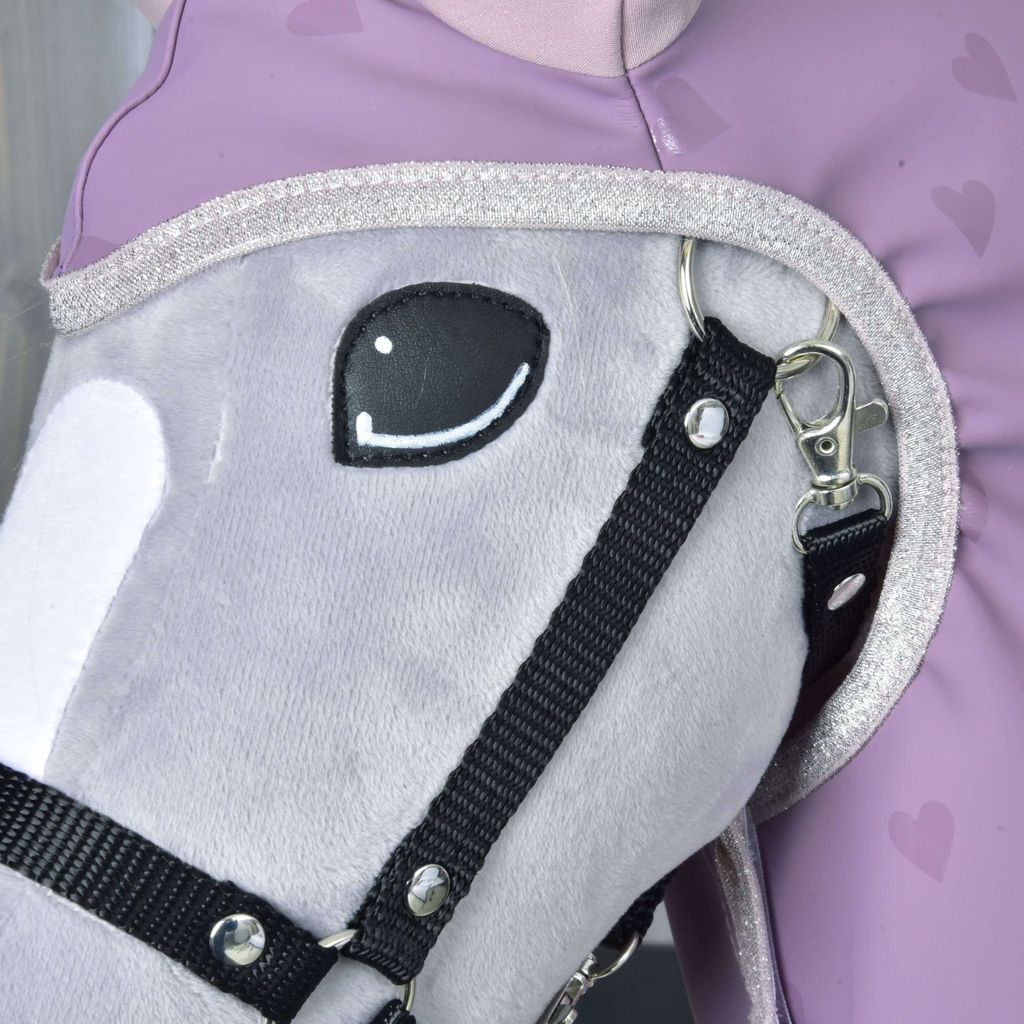 Close-up van de pluchen grijze Hobby Horse van Hobby Horse LarDen met zwart hoofdstel, die het accessoire Regenjas Sweetheart draagt, naast een persoon in een lavendelkleurige outfit met hartpatroon. Het paard heeft een geborduurd glimlachend oog en glinsterende bies langs de hals.