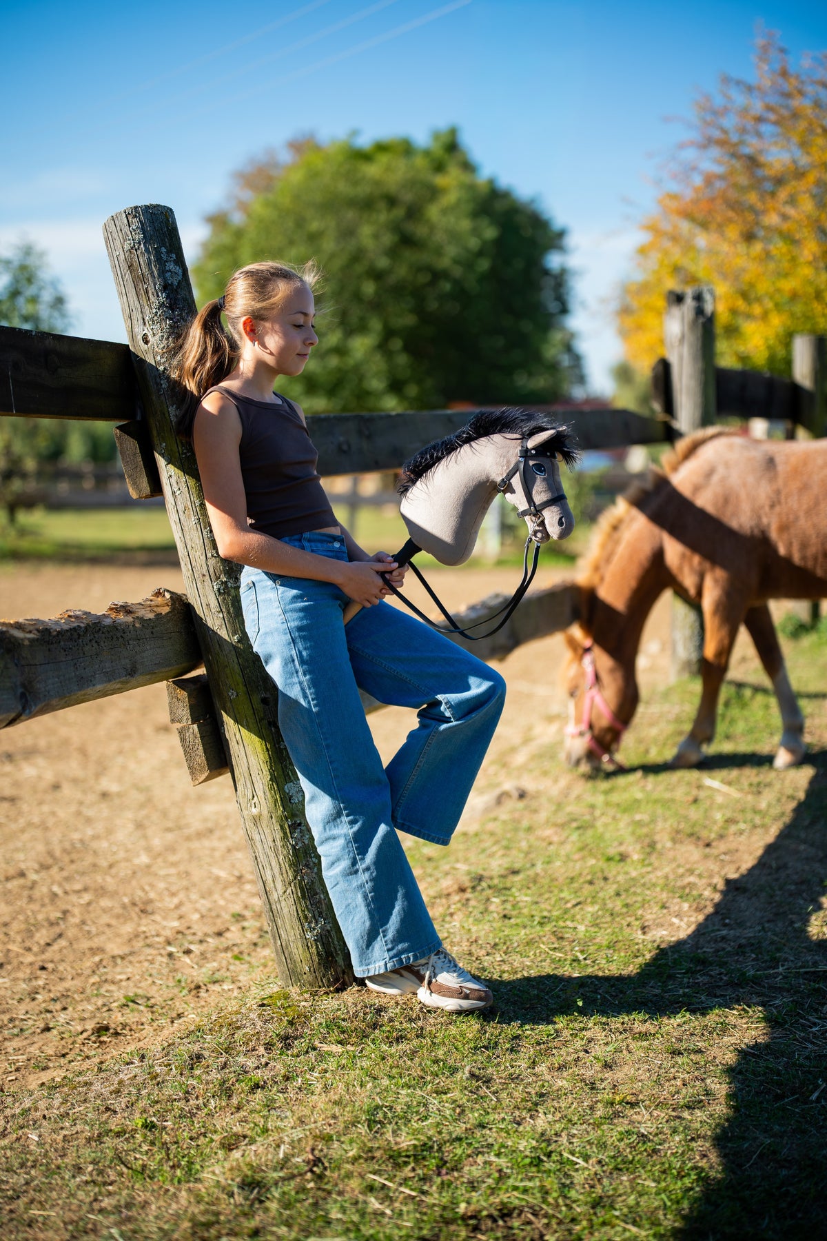 Meisje in een bruine top en jeans leunt tegen een houten hekpalen met Hobby Horse Thor met zwart Halster van Hobby Horse LarDen, grijze pluchen kop met zwarte garenmane, met een grazende pony in de paddock en het Complete zwarte hoofdstel met diamanten