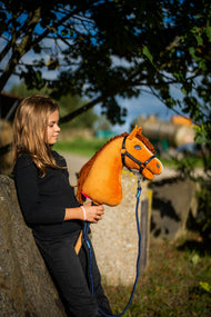 Lifestylefoto: een meisje in een zwarte top leunt onder een boom tegen een stenen blok met Hobby Horse Wendy met zwart Halster van Hobby Horse LarDen – helder voskleurige pluchen kop, gemberkleurige manen van garen – hier getoond met het Blues rope stripe-halstertouw