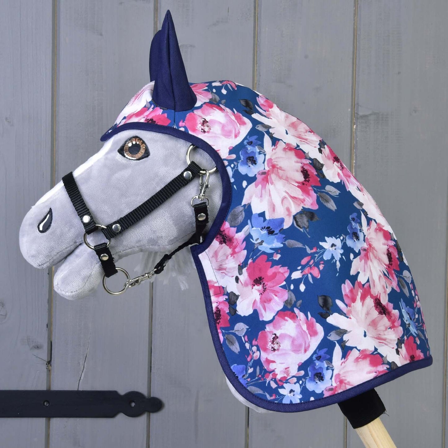 Regenjas Pioenroos van Hobby Horse LarDen, een grijze Hobby Horse met een zwart hoofdstel, die een blauwe regenjas met roze en witte bloemen draagt, gemonteerd op een houten stok voor een grijze houten wand.